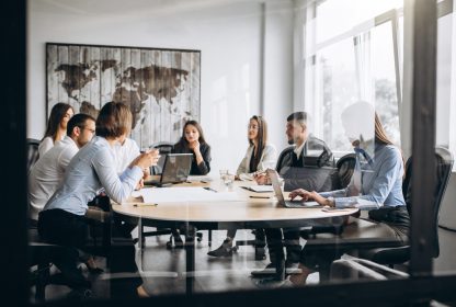 Lean Office: Pessoas com roupas formais sentadas em uma mesa oval, realizando uma reunião.