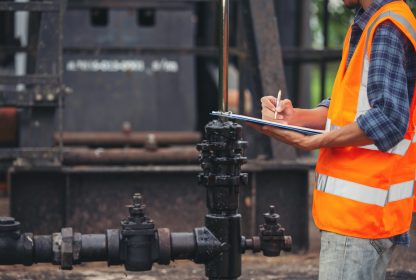 Análise preliminar de riscos: trabalhador em pé checando sua prancheta enquanto máquinas trabalham.