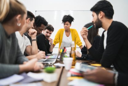 Controle de tarefas: mulher negra ao fundo da imagem olhando para um mesa com pessoas sentadas dos dois lados.
