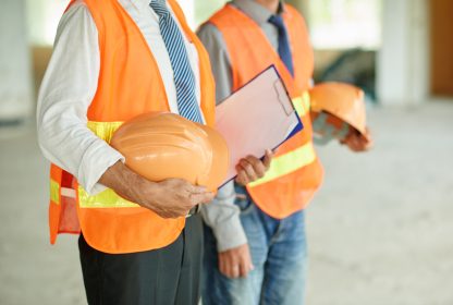 Dois homens com coletes de segurança do trabalho, capacetes e uma prancheta nas mãos