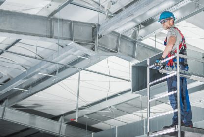 Instalador de circulação de ar de armazém em andaimes de alumínio se preparando para fazer os ajustes finais das saídas de ar.